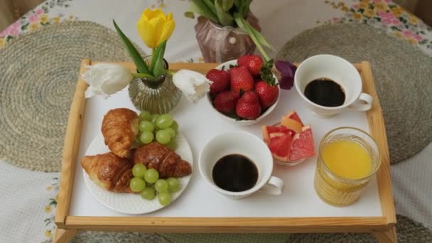 Närbild av en trä bricka med varmt kaffe, apelsinjuice, frukt croissant och blommor. — Stockvideo