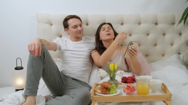 Bella coppia attraente prendendo selfie durante la colazione nel letto, sdraiato a letto. — Video Stock