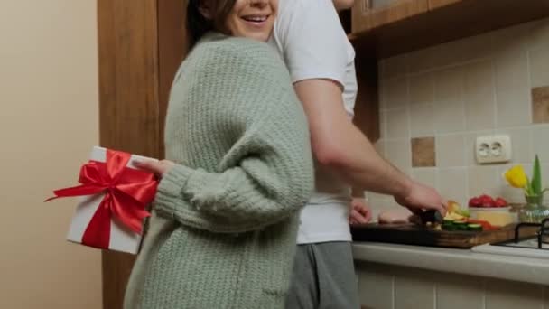 Młoda kobieta daje prezent mężowi, który przygotowuje jedzenie w kuchni. — Wideo stockowe
