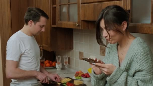 Young man prepares food in the kitchen while his wife is surf the internet. — Stock Video