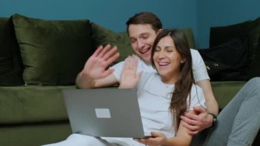 Dost canlısı genç bir çift, akrabalarıyla görüntülü görüşme yapıyor, dizüstü bilgisayarlarına el sallıyorlar..