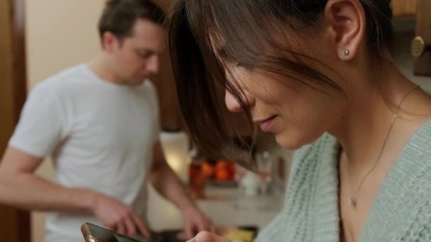 Unge man lagar mat i köket medan hans fru surfar på internet. — Stockvideo