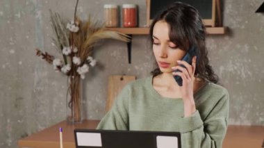 Oturma odasında dizüstü bilgisayarda oturan, telefonla konuşan ve notlar yazan bir kız..