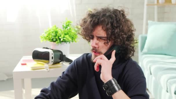 Jovem de cabelos encaracolados homem falando ao telefone enquanto sentado no chão perto do sofá. — Vídeo de Stock