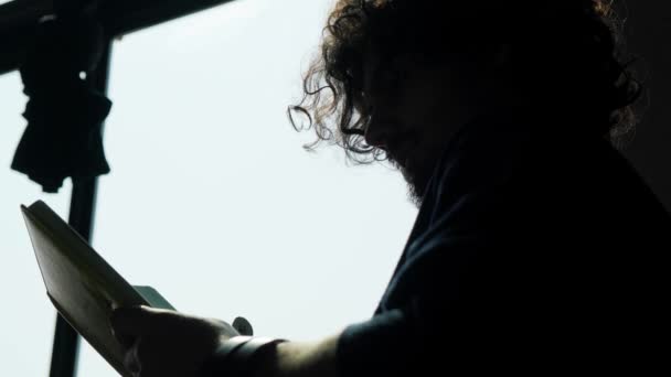Η σιλουέτα ενός ανθρώπου που κάθεται κοντά στο παράθυρο και διαβάζει ένα βιβλίο στο ηλιοβασίλεμα. — Αρχείο Βίντεο