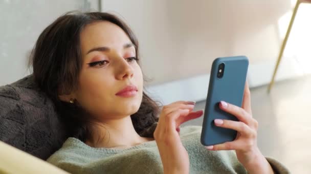 Femeia navighează pe internet pe telefonul mobil în timp ce se relaxează pe o canapea în camera ei de zi . — Videoclip de stoc