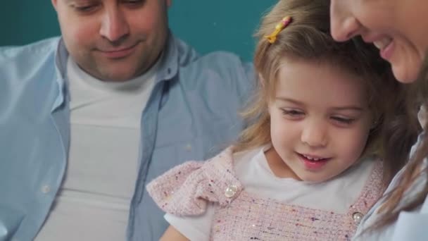 Jonge familie leest een boek voor aan hun dochtertje terwijl ze op de bank zitten. — Stockvideo