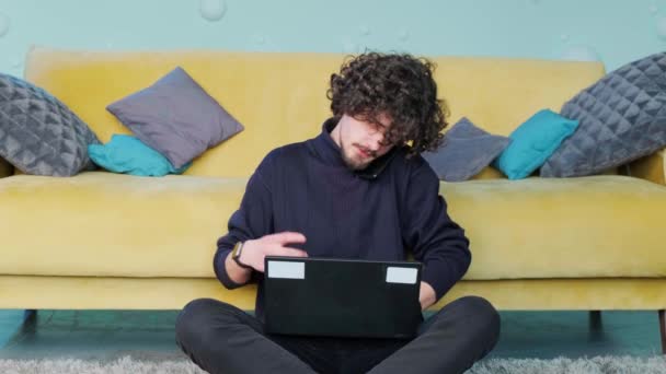 Jongeman praat aan de telefoon en werkt aan de computer terwijl hij op de vloer zit. — Stockvideo