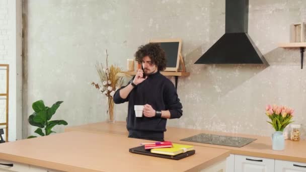 Egy göndör hajú fiatalember beszél telefonon, miközben egy csésze teát iszik.. — Stock videók