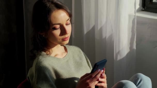 Bella donna seduta vicino alla finestra e utilizzando il telefono cellulare al tramonto. — Video Stock