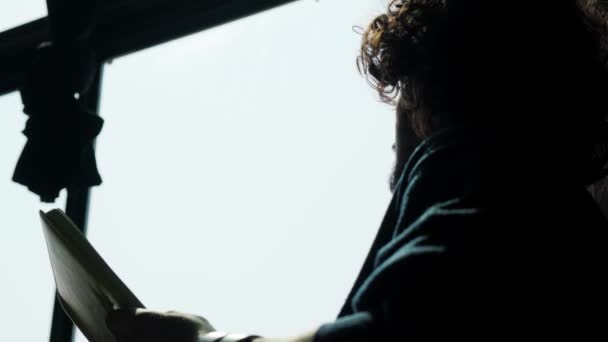窓の近くに座って日没時に本を読んでいる男のシルエット. — ストック動画