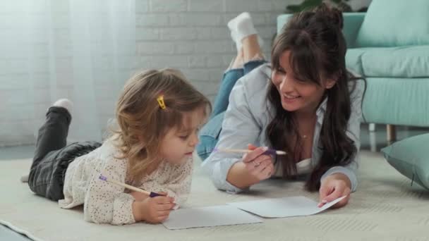 Anya és lánya a szőnyegen fekszenek a földön, együtt rajzolnak papíron.. — Stock videók