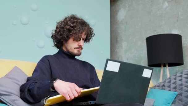 A fiatalember a laptopját nézegeti, és a jegyzetfüzetébe ír.. — Stock videók
