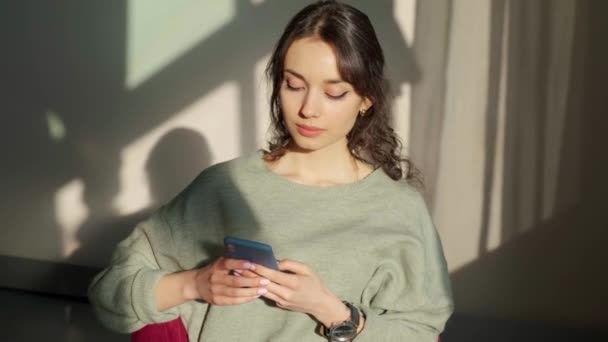 Piękna kobieta siedzi w pobliżu okna i używa telefonu komórkowego o zachodzie słońca. — Wideo stockowe