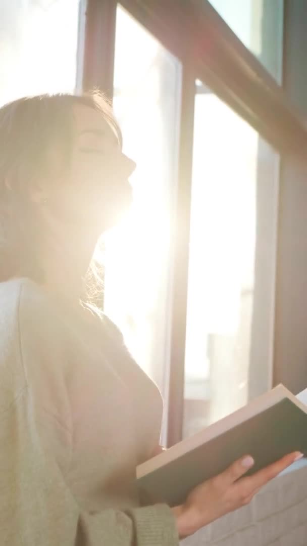 Krásná žena sedí v blízkosti okna a čtení knihy na pozadí západu slunce. — Stock video