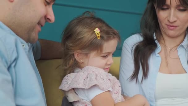 幸せな家族はソファに座っている間に彼らの小さな娘に本を読む. — ストック動画