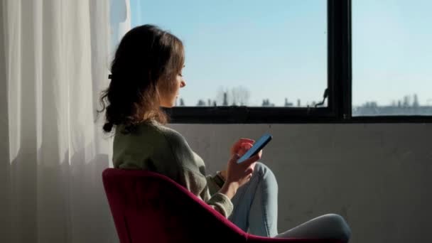 Belle femme assise près de la fenêtre et utilisant un téléphone mobile au coucher du soleil. — Video