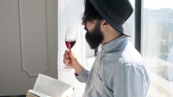Zadowolony brodaty mężczyzna pijący czerwone wino podczas czytania książki w domu. — Wideo stockowe