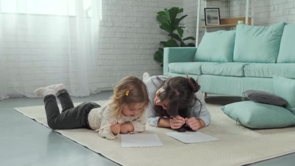 Anya és lánya a szőnyegen fekszenek a földön, együtt rajzolnak papíron.. — Stock videók