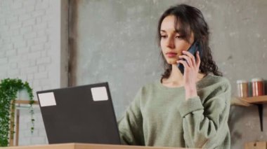 Oturma odasında dizüstü bilgisayarda oturan, telefonla konuşan ve notlar yazan bir kız..