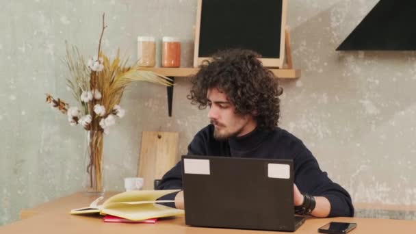 Ernstige krullenbol jongeman werken op de computer tijdens het zitten in de keuken. — Stockvideo