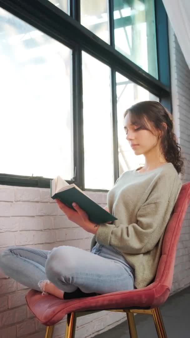 Красивая женщина сидит у окна и читает книгу на фоне восхода солнца. — стоковое видео