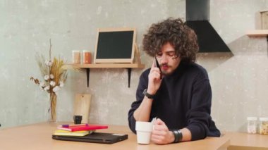 Mutfaktaki adam gülümsüyor, kahve içiyor ve telefonda konuşuyor..