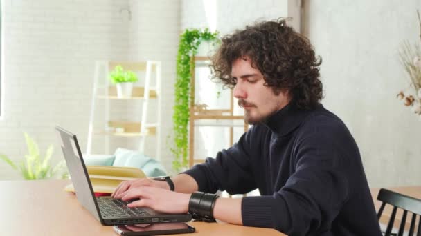 Vážné kudrnaté vlasy mladý muž pracující na počítači, zatímco sedí v kuchyni. — Stock video