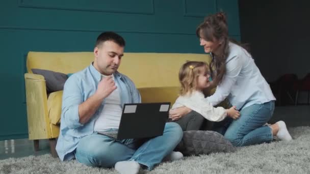 Kislány játszik anyával a szőnyegen, míg az apja számítógépet használ.. — Stock videók