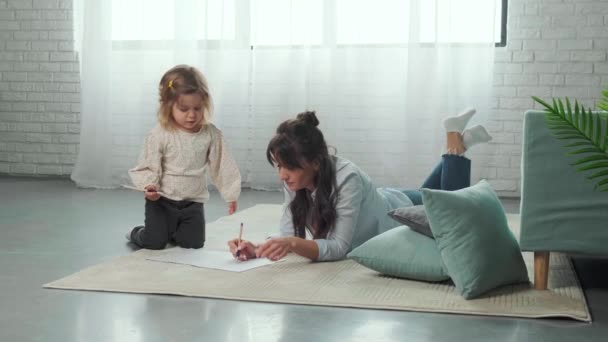 Mamá e hija tumbadas en la alfombra en el suelo, dibujando sobre papel juntas. — Vídeos de Stock