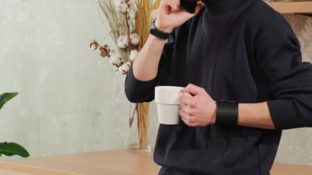 A férfi a konyhában mosolyog, kávét iszik és okostelefonon beszél.. — Stock videók