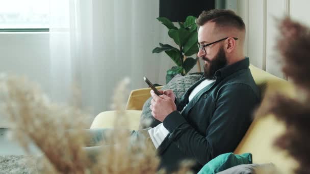 Bearded man sitter i vardagsrummet på soffan hemma chatta, skriva meddelande med hjälp av cellulära. Affärsman hemma löser affärsärenden per telefon. Ny generation beroende av enheter koncept — Stockvideo