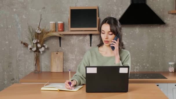 Meisje zit achter laptop in haar woonkamer, telefoneert en schrijft notities. — Stockvideo