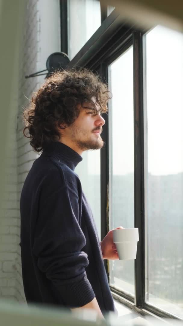 Muž s kudrnatými vlasy, dívající se z okna, přemýšlející, pijící šálek čaje nebo kávy. — Stock video