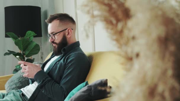 千年紀の髭を生やした男が家のチャットでソファに座って携帯電話でメッセージを入力して. — ストック動画