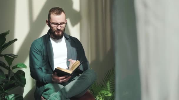 Joven barbudo sentado cerca de la ventana y leyendo un libro al amanecer. — Vídeo de stock