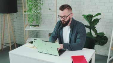Sakallı adam dizüstü bilgisayara yazar ve not defterine not yazar..