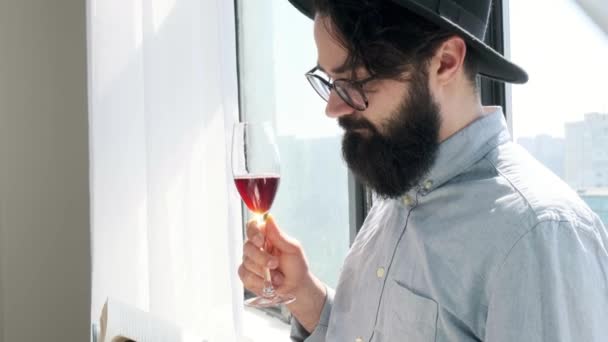 Homem barbudo agradável bebendo vinho tinto enquanto lê um livro em casa. — Vídeo de Stock
