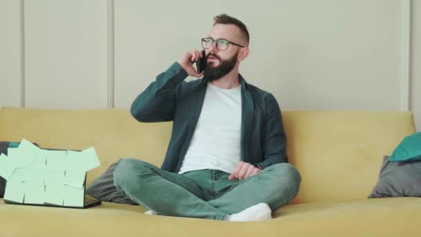 Cieszymy się wolnym dniem w domu. Radosny brodaty młodzieniec rozmawiający przez telefon komórkowy. — Wideo stockowe