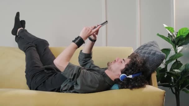 Curly cabelo jovem no sofá ouvindo música com fones de ouvido. — Vídeo de Stock
