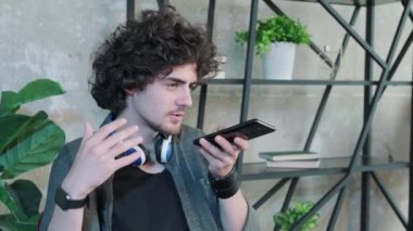 Kıvırcık saçlı adam ofisteki masada oturur ve akıllı telefondan hoparlörle konuşur..