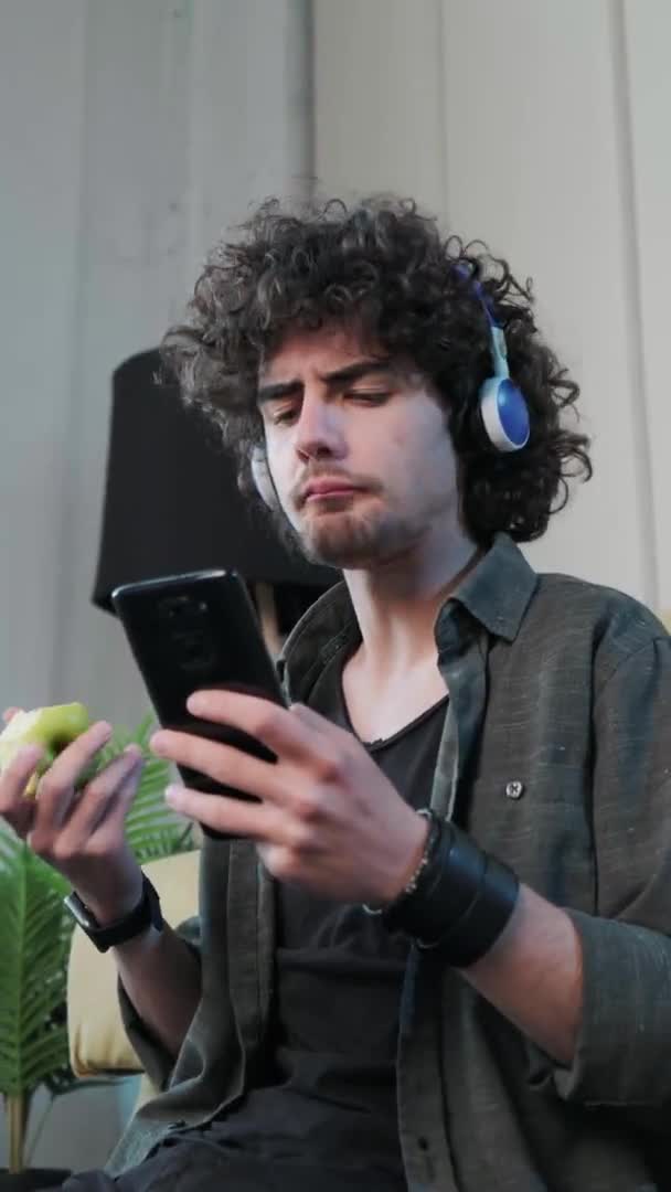 Young man listening to the music and gesturing while eatting an apple, vertical video. — Stock Video