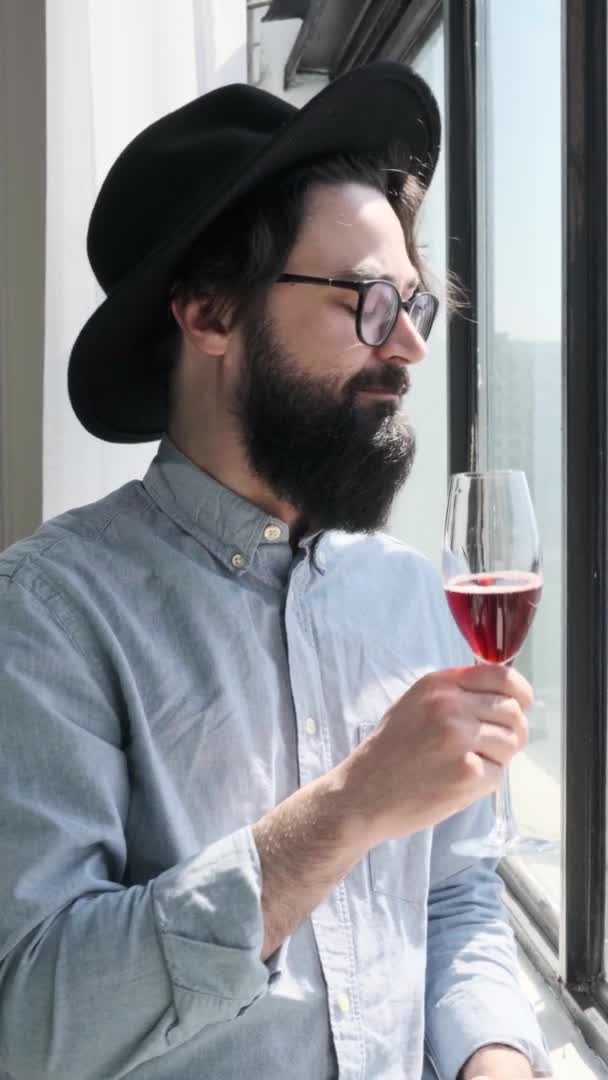 Pleased bearded man drinking red wine and admiring view form window at home. — Stock Video