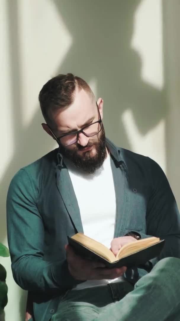 Szakállas fiatalember ül az ablak mellett, és könyvet olvas, függőleges videó. — Stock videók