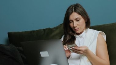 Genç ciddi bir kadın dizüstü bilgisayarla kanepede uzanıyor, cep telefonu kullanıyor ve çalışıyor..