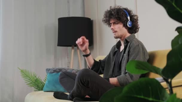 卷发年轻人坐在沙发上，用耳机听音乐. — 图库视频影像