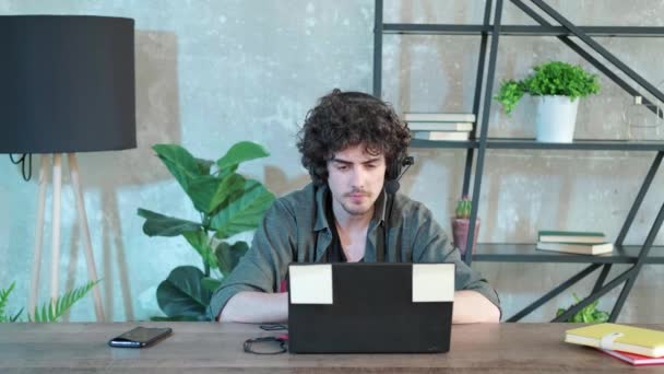 Giovane uomo dai capelli ricci utilizzando computer portatile con auricolare per parlare online. — Video Stock