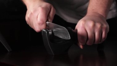 Bir adam bileyiciyle bıçağı keskinleştirir. Karanlık bir arka planda eylem. & Kapat.