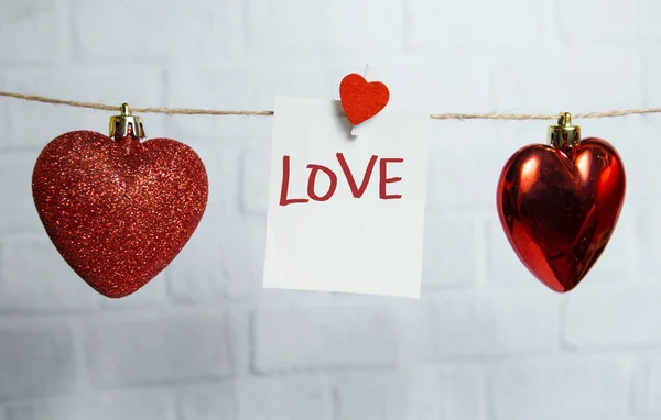 Red hearts and white card hang on a thread on a white brick background — Stock Photo, Image