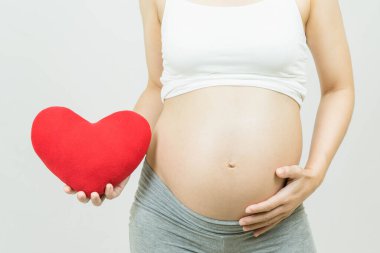 Elinde kırmızı bir kalbi olan hamile bir kadının seçici odak noktası gri arka plan, vücut parçası, mutlu hamilelik zamanı, yeni yaşam konsepti.
